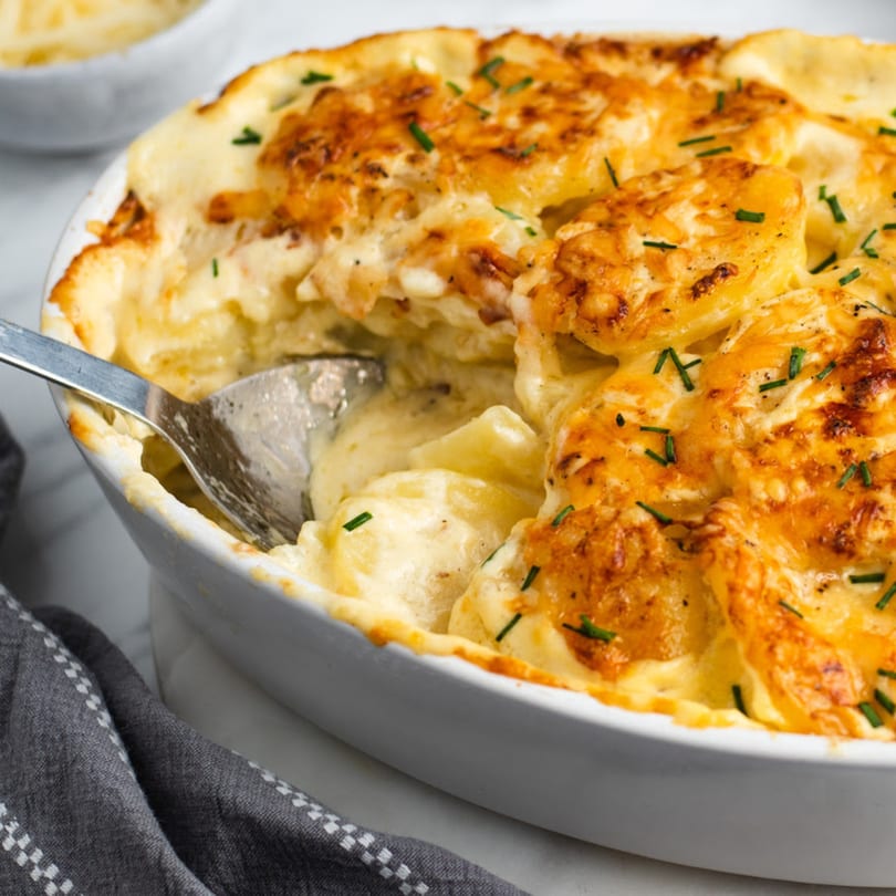 Close-up of golden-brown scalloped potatoes with chives in a white baking dish, with a spoon.