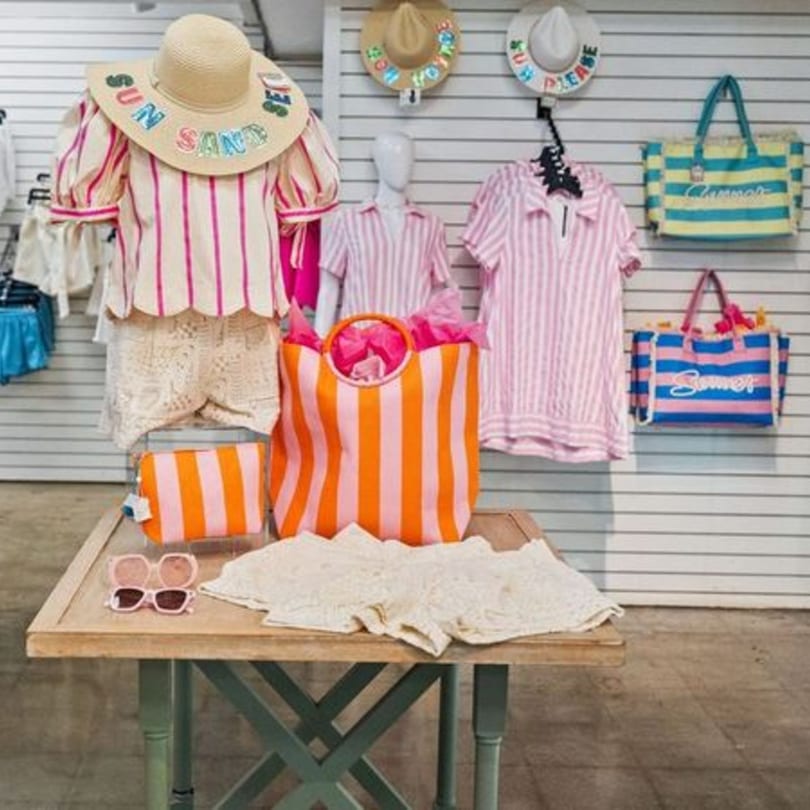 A retail display features summer clothing, hats, and bags in pink, orange, and blue stripes.