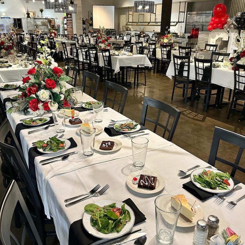A dining room set for an event with tables draped in white, black chairs, and floral centerpieces.