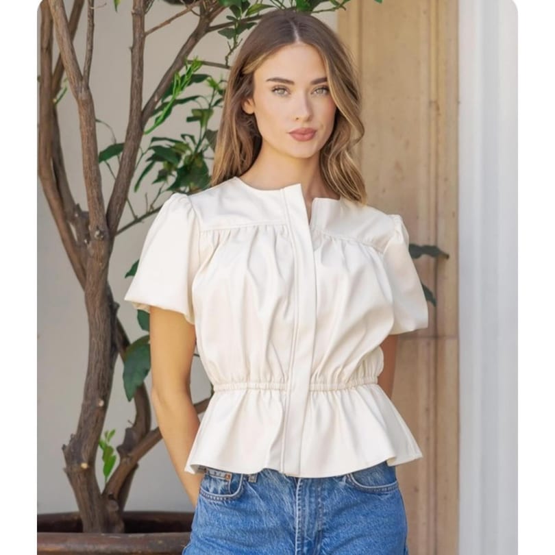 A woman with long brown hair wearing a cream-colored top and blue jeans stands by a potted plant.