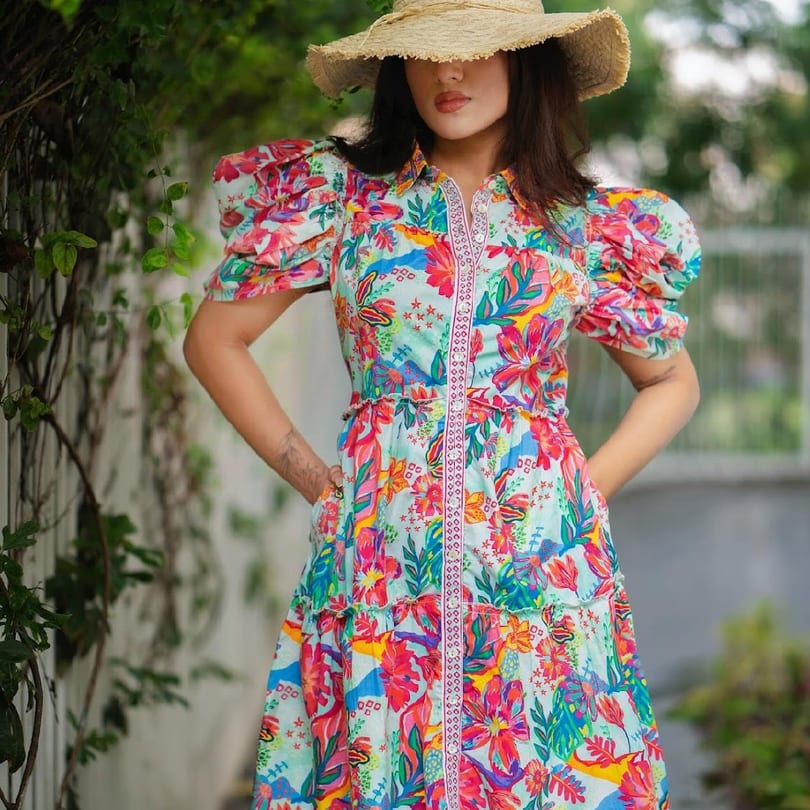 A woman in a colorful floral dress and straw hat stands outdoors with hands in pockets.