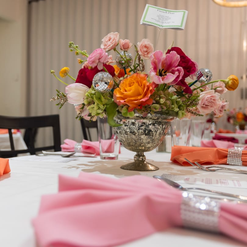 A vibrant floral centerpiece with disco ball accents on a white tablecloth with pink and orange napkins.