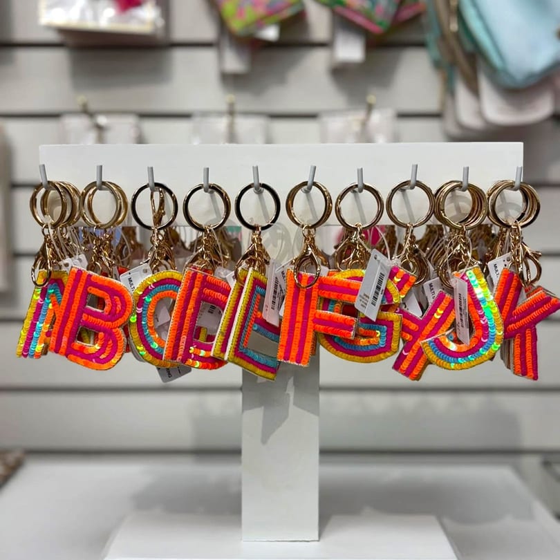 Colorful, sequined letter keychains, spelling out "ABCDEFGHIJ", hang on a white display stand.