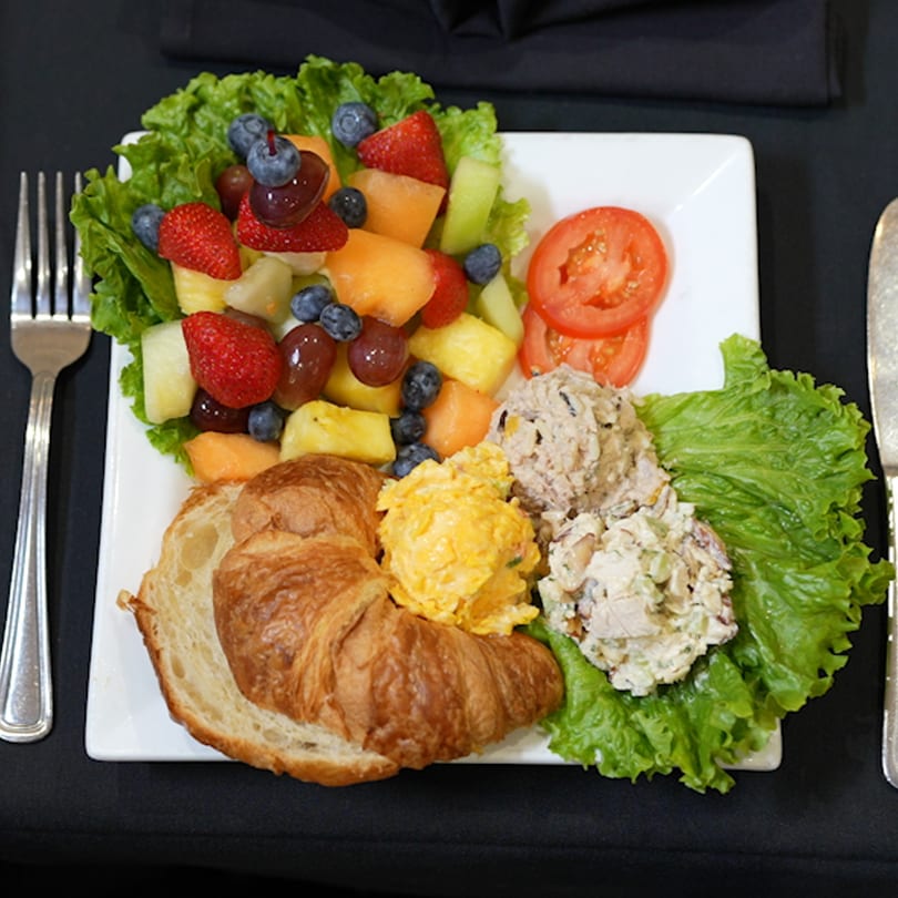 A plate with a croissant, fruit salad, tomato slices, and two scoops of salad on lettuce.